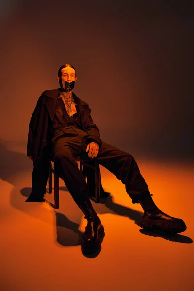 Jovem bonito em roupa elegante com máscara amarrada sentado relaxantemente na cadeira, conceito de moda — Fotografia de Stock