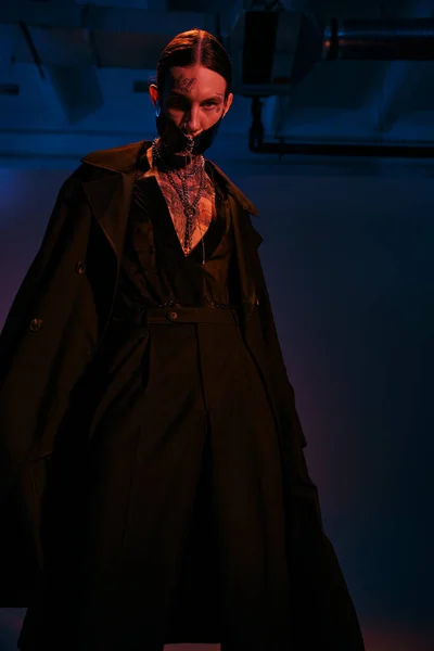 Tiro vertical de hombre joven con estilo en traje futurista negro posando en luces oscuras, concepto de moda - foto de stock