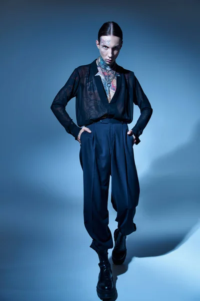 Молодой симпатичный мужчина в стильной черной одежде с татуировками с руками в карманах, смотрящий в камеру — стоковое фото