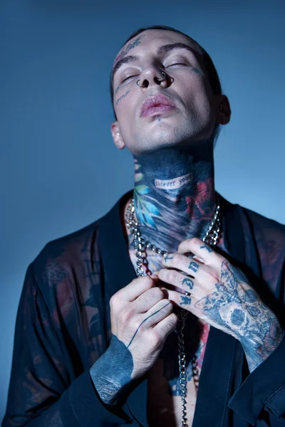 Handsome alluring man with stylish tattoos posing with closed eyes touching his accessories, fashion — Stock Photo