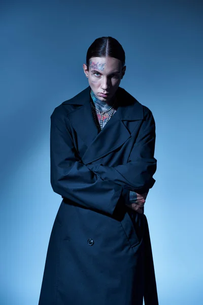 Handsome young man with tattoos and piercing wrapping in his black coat looking at camera, fashion — Stock Photo