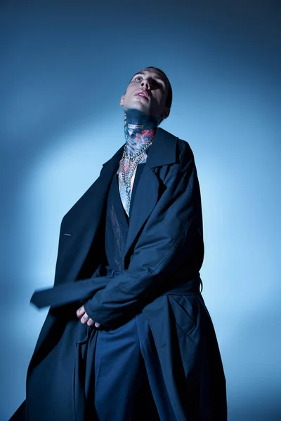 Вертикальний знімок сексуального молодого чоловіка з татуюваннями та аксесуарами, що позують у чорному пальто та дивляться вгору — стокове фото