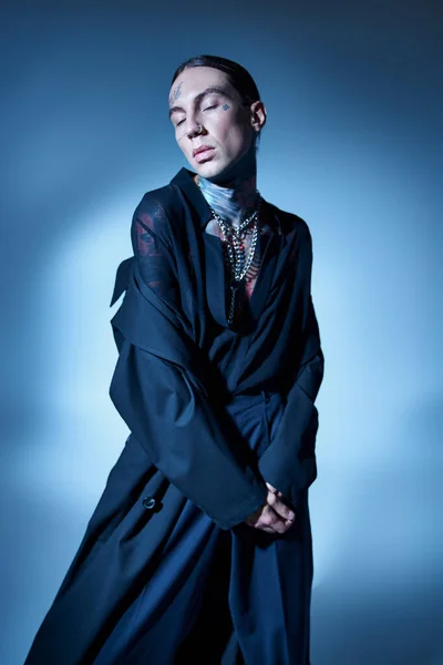 Tiro vertical de hombre joven con estilo con tatuajes con los ojos cerrados en abrigo negro, concepto de moda - foto de stock