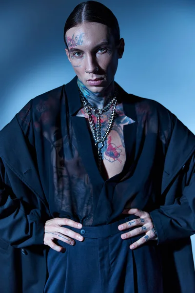 Atractivo seductor hombre con tatuajes en traje negro posando con las manos en las caderas mirando a la cámara - foto de stock
