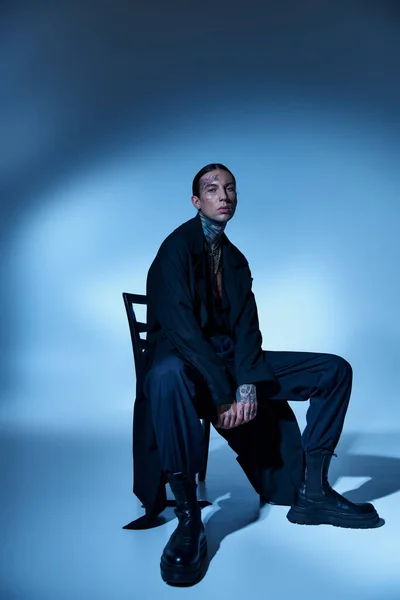 Hombre elegante guapo en traje voguish negro sentado en la silla y mirando a la cámara, concepto de moda - foto de stock