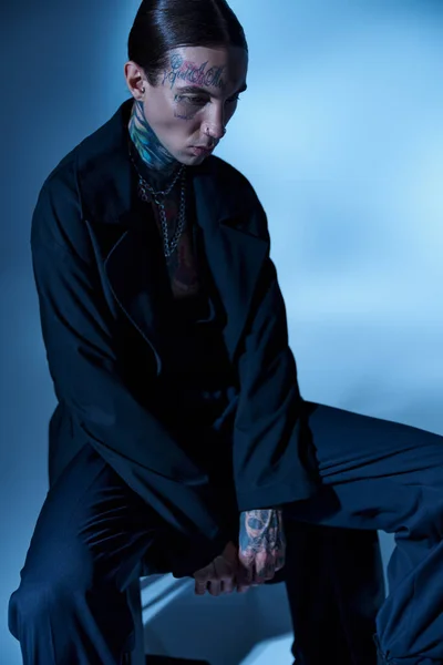 Tiro vertical de belo jovem sentado na cadeira em traje preto e olhando para longe, moda — Fotografia de Stock
