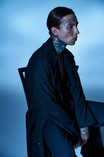 Hombre atractivo con estilo en abrigo negro con tatuajes sentados en la silla y mirando hacia otro lado, concepto de moda - foto de stock