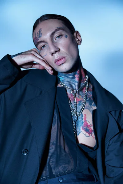 Portrait of alluring voguish man with tattoos and piercing looking at camera with hand near face — Stock Photo