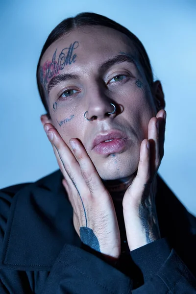 Retrato de homem bonito elegante com tatuagens no rosto olhando para a câmera com as mãos no rosto, moda — Fotografia de Stock
