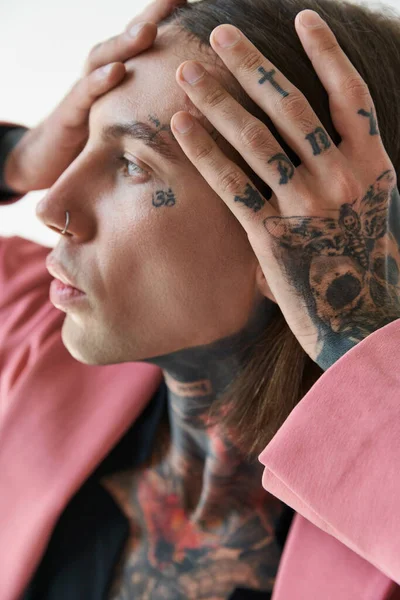 Retrato de hombre joven de moda con tatuajes en la cara y piercing con las manos en la cara mirando hacia otro lado - foto de stock