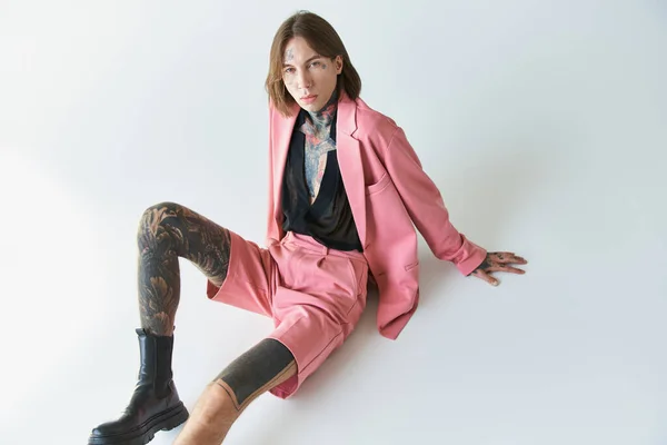 Jovem sedutor homem com tatuagens em traje rosa sentado no chão olhando para a câmera, conceito de moda — Fotografia de Stock