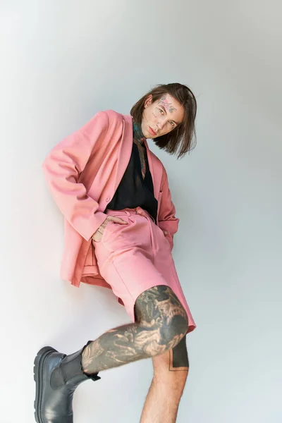 Good looking sexy man with tattoos in stylish outfit posing with leg on wall and hands in pockets — стоковое фото