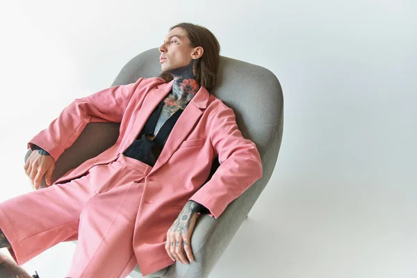 Sexy junger Mann in stylischem rosa Outfit entspannt auf bequemem Stuhl und schaut weg, Modekonzept — Stockfoto