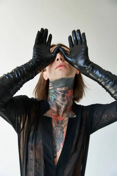 Young alluring man with tattoos and piercing with hands in leather gloves on face, fashion concept — Stock Photo