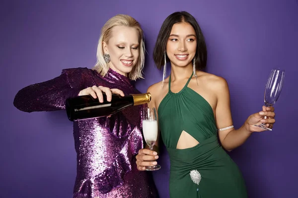 Happy woman pouring champagne into glass of asian female friend on purple backdrop, New Year 2024 — Stock Photo