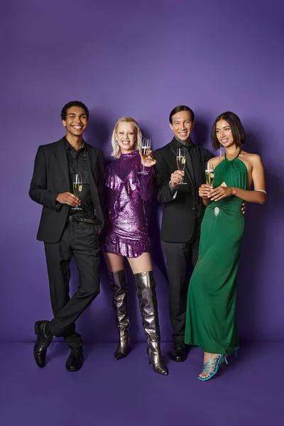 Fiesta de Navidad, felices amigos multiétnicos en traje festivo sosteniendo copas de champán en púrpura - foto de stock