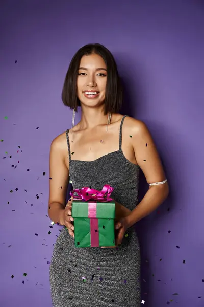 Radioso asiatico donna in partito abito holding avvolto Natale regalo vicino cadere coriandoli su viola — Foto stock