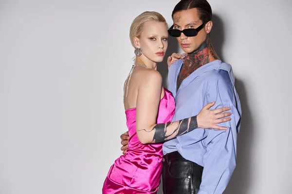 Блондинка в розовом платье обнимается с татуированным парнем в солнечных очках на сером, Новый 2024 год — стоковое фото