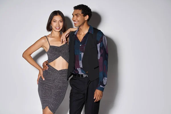 Cheerful asian woman in party dress posing with african american man on grey, Christmas concept — Stock Photo