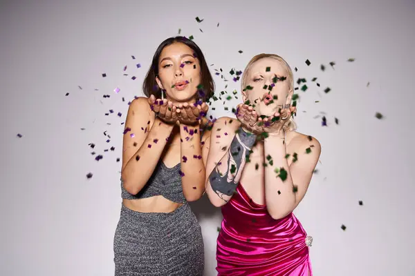 Duas mulheres multiétnicas em vestidos de festa soprando confete em fundo cinza, conceito de festa de Ano Novo — Fotografia de Stock