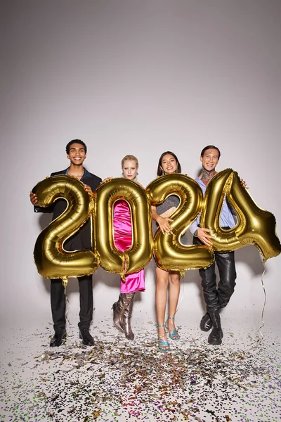 Four happy interracial friends holding balloons with 2024 numbers near confetti, New Year party — Stock Photo