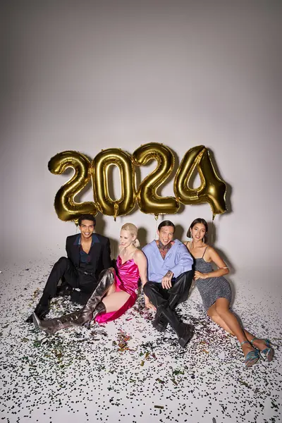 Groupe d'amis multiethniques assis près de confettis et ballons avec 2024 numéros, Bonne année — Photo de stock