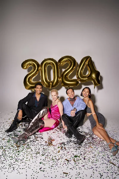Persone multiculturali sorridenti sedute vicino a coriandoli e palloncini con 2024 numeri, felice anno nuovo — Foto stock