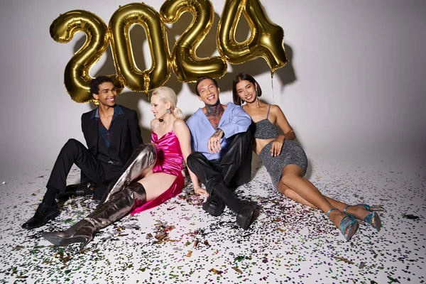 Happy multicultural people sitting near confetti and balloons with 2024 numbers, Happy New Year — Stock Photo