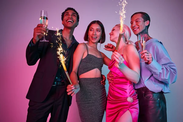 New Year celebration, joyful four multiethnic friends holding sparklers and champagne on purple pink — Stock Photo