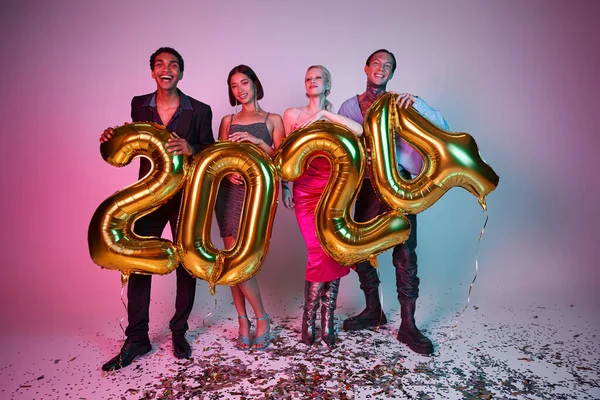 Feliz Año Nuevo, felices amigos multiétnicos con atuendo festivo sosteniendo globos con números 2024 - foto de stock