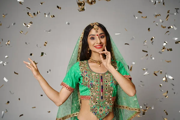 Alegre mujer india joven en ropa nacional con bindi y velo verde rodeado de confeti - foto de stock