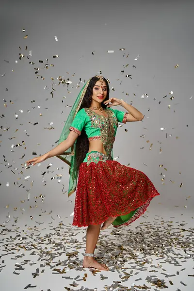 Tiro vertical de mujer india joven con bindi de pie sobre una pierna y gesto bajo la lluvia confeti - foto de stock