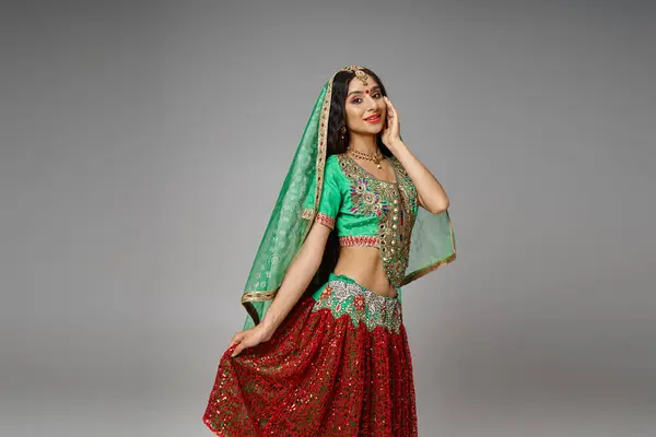 Alegre mujer india en verde choli tocando el dobladillo de su falda roja posando con la mano en la mejilla - foto de stock