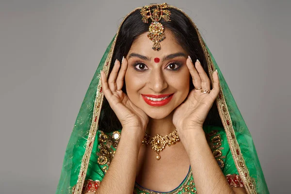 Joyeuse jeune femme indienne avec point bindi et voile vert souriant à la caméra avec les mains près du visage — Photo de stock