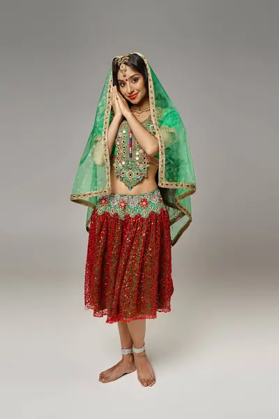 Alegre mujer india en falda roja y verde choli posando sobre fondo gris y mirando a la cámara - foto de stock