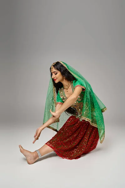 Mulher indiana atraente em traje nacional com bindi dot gesto enquanto dança em pano de fundo cinza — Fotografia de Stock