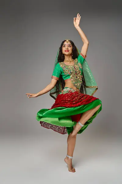 Mujer india joven en traje nacional de pie en una pierna con el brazo levantado mientras baila activamente - foto de stock
