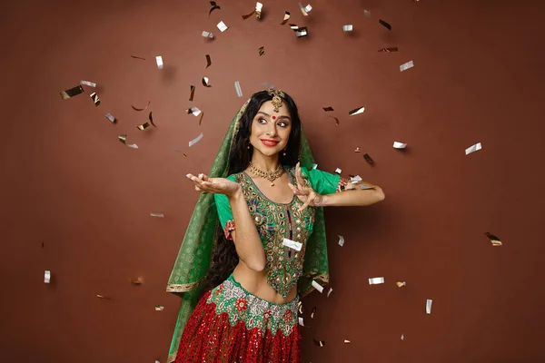 Joyous indian woman in national attire with bindi gesturing while dancing under confetti rain — Stock Photo