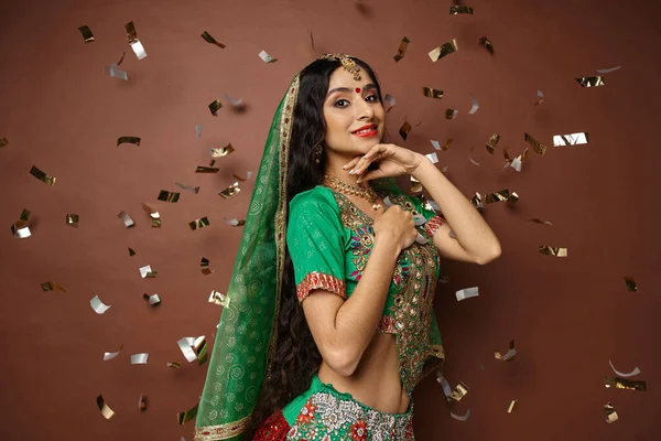 Bonita mujer india con punto bindi y velo verde posando bajo la lluvia confeti con la mano bajo la barbilla - foto de stock