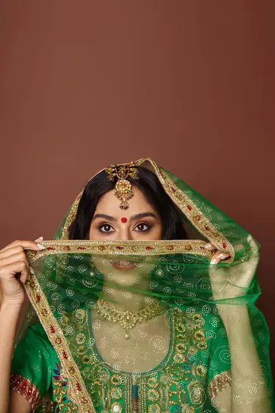 Tiro vertical de mujer india joven en traje nacional con bindi cubriendo su as con velo verde - foto de stock