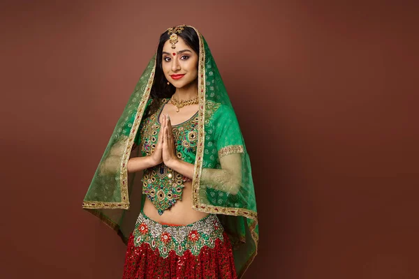 Jolie femme indienne joyeuse avec bindi et voile vert montrant geste de prière et souriant à la caméra — Photo de stock