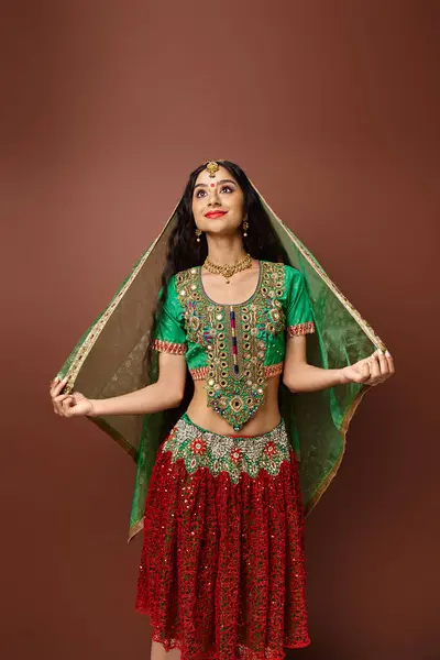 Vertikale Aufnahme einer attraktiven, fröhlichen Indianerin, die vor braunem Hintergrund posiert und ihren grünen Schleier berührt — Stockfoto