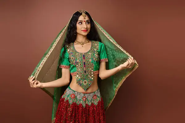 Hermosa mujer india en traje nacional tocando el velo verde y mirando hacia otro lado en el fondo marrón - foto de stock