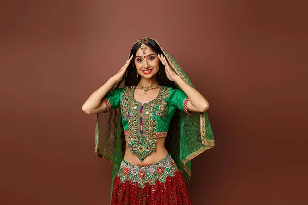Atractiva mujer india joven en ropa tradicional posando con las manos cerca de la cara sobre el telón de fondo marrón - foto de stock