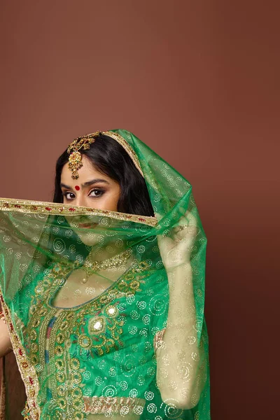 Plan vertical de jeune femme indienne couvrant son visage avec un voile vert posant sur fond brun — Photo de stock