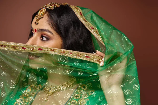 Привлекательная индийская женщина с длинными волосами и точкой бинди, закрывающая лицо зеленой вуалью, отворачивающаяся — стоковое фото