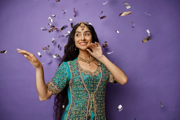 Mujer india alegre en ropa nacional con accesorios sonriendo y posando bajo la lluvia confeti - foto de stock