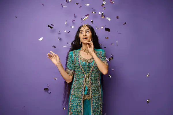 Hermosa mujer india asombrada posando bajo la lluvia confeti sobre fondo púrpura con la mano cerca de la cara - foto de stock