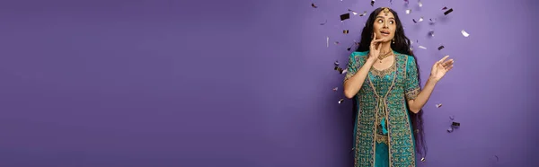 Surprised young indian woman under confetti rain on purple backdrop with hand near face, banner — Stock Photo