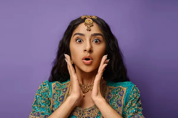 Belle femme indienne choquée posant sur fond violet avec les mains près du visage en regardant la caméra — Photo de stock